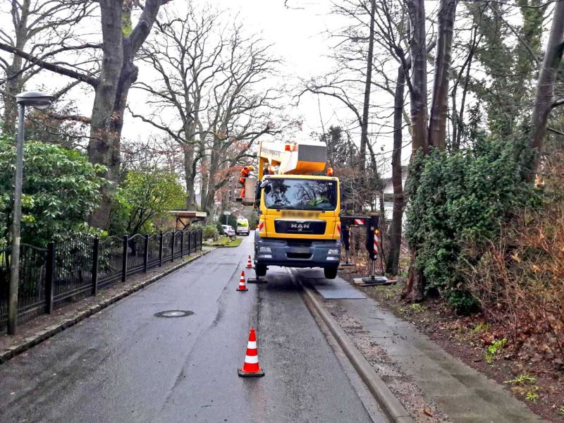 LKW-Kran mit seitlichen Stützen auf einer engen Straße, abgesichert mit Pylonen. Links und rechts der Straße stehen hohe, kahle Bäume.