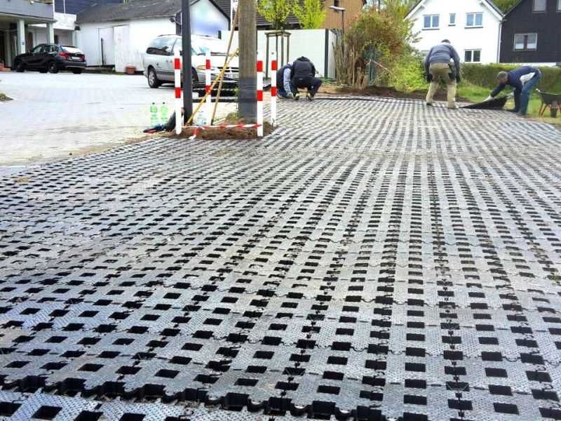 Mehrere Personen legen eine Fläche aus Rasengittersteinen an. Links davon sind eine Straße und Gebäude sichtbar.