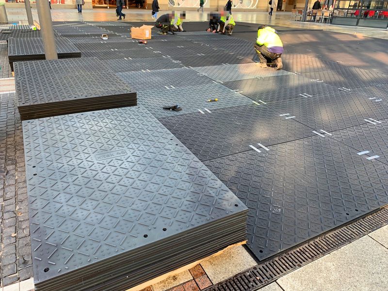 Bauarbeiter verlegen die x:tek46 Bodenschutzplatten als Fläche in einer Fußgängerzone. Im Hintergrund sieht man Ladenfronten.