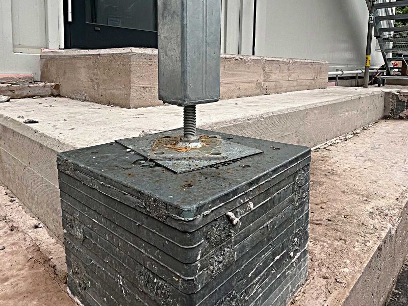 Mehrere JakStak Unterlegplatten wurden auf einer Treppenstufe aus Beton gestapelt, um eine stabile Unterlage für den Metalfuß einer Treppe zu bieten. Die Treppe steht vor weißen Raumcontainern und dient als Zugang zu der oberen Reihe der Container.