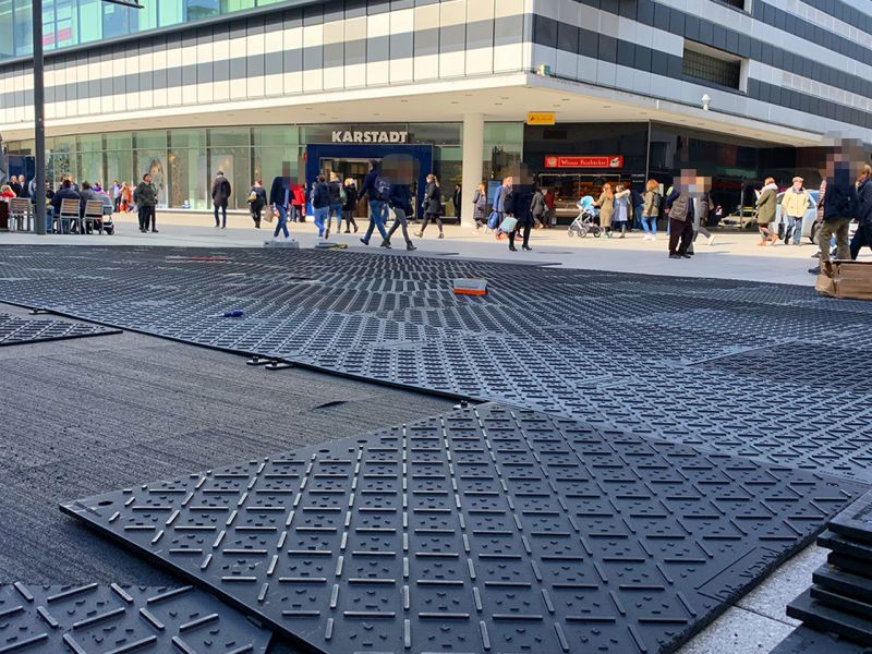Bild von der Frankfurter Zeil. Im Vordergrund liegen Kunststoff-Fahrplatten auf dem Boden. Im Hintergrund sieht man ein großes Gebäude, ein Kaufhaus. Davor laufen viele Passanten vorbei.