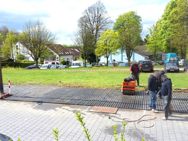 Neben einer gepflasterten Straße wurde mit Rasengittersteinen aus Kunststoff ein Parkplatz angelegt. Man sieht Bauarbeiter und eine kleine Walze. Im Hintergrund sind Bäume und Gebäude.