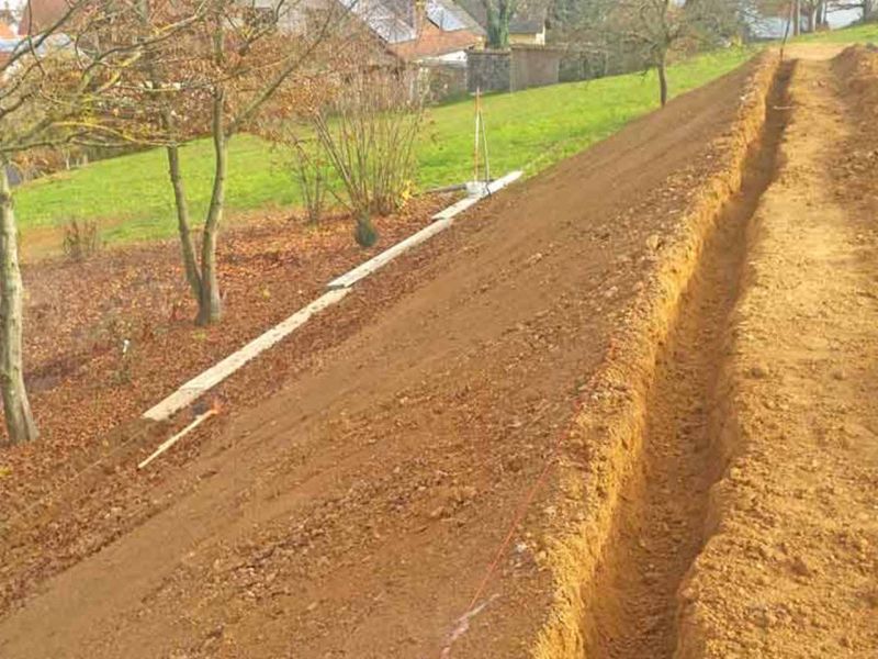 Erdarbeiten an künstlich angelegtem Hang. Ein Graben verläuft am oberen Rand. Am Fuß des Hangs liegen Bretter. Bäume und Häuser im Hintergrund.