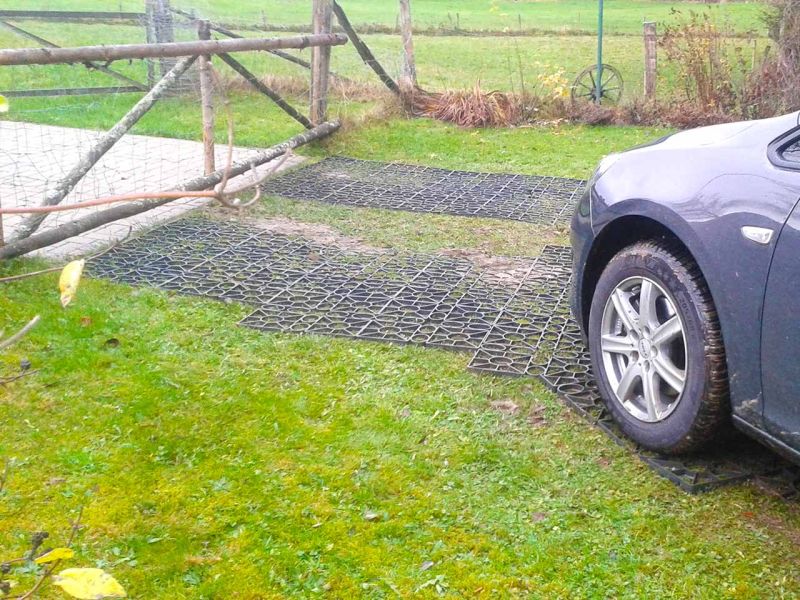 Auf einer Rasenfläche liegen Rasengitter, die eine Fahrstraße bilden. Der Weg endet an einem Gatter. Man sieht einen Teil eines darüberfahrenden Autos.