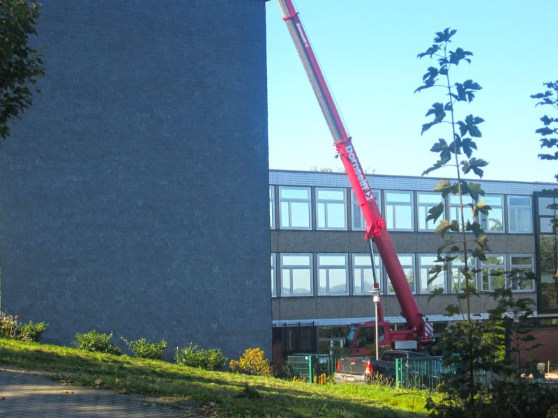 Mobilkran hebt Bauteil an Schulfassade ein und zeigt sicheren Einsatz dank stabiler Arbeitsplattform.