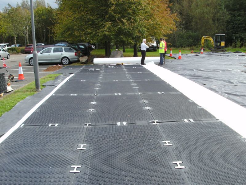 Zwei Personen bei der Verlegung einer schwarzen Plattenfläche neben geparkten Autos und Baumaschinen.