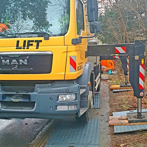 Gelber LKW-Kran mit ausgefahrenen Stützen auf Kunststoffplatten zur Lastverteilung. Rechts davon stehen Bäume.