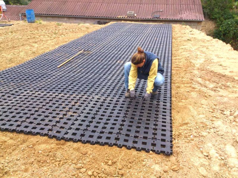 Eine Frau verlegt Rasengittersteine auf einer erdigen Fläche. Im Hintergrund ist das Dach einer Halle zu sehen. Ein Rechen liegt auf dem Boden.