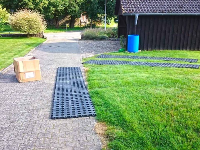 Kunststoff-Rasengitter RGS35 auf Gehweg und Rasen ausgelegt. Karton und blaues Fass daneben.