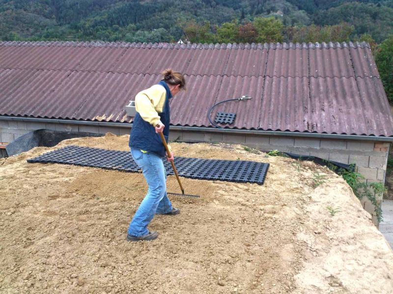 Eine Frau in blauer Weste und heller Jacke bearbeitet mit einer Harke eine sandige Fläche. Sie steht auf einer leicht erhöhten, erdigen Böschung. Auf dem Boden liegen schwarze Kunststoff-Rasengitterplatten, die offenbar verlegt werden sollen. Im Hintergrund sind grüne Bäume und ein bewaldeter Hügel zu sehen.