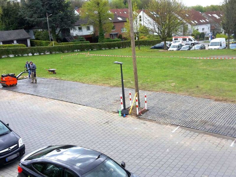 Ein Parkplatz aus Rasengittersteinen, der auf einer Wiese neben einer Straße angelegt wurde. Man sieht verschiedene Fahrzeug und Gebäude im Hintergrund.