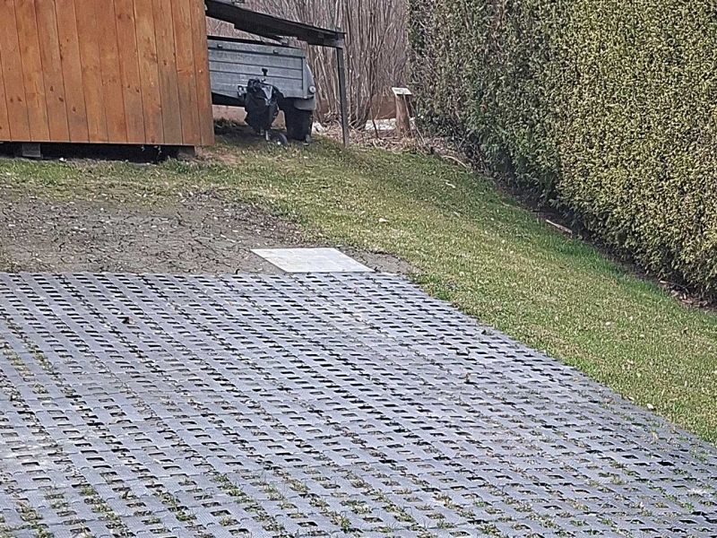 Auf einem Rasen wurden Kunststoff-Rasengittersteinen als Fläche verlegt. Rechts eine Hecke, im Hintergrund einen Anhänger neben einem Holzschuppen.