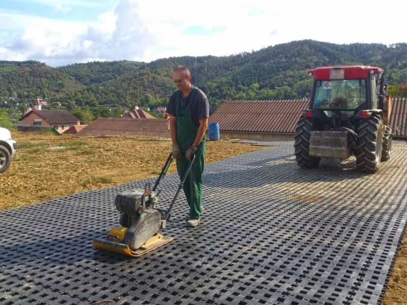 Ein Mann schiebt eine Rüttelplatte über frisch verlegte Rasengittersteine aus Kunststoff. Dahinter steht ein Traktor.