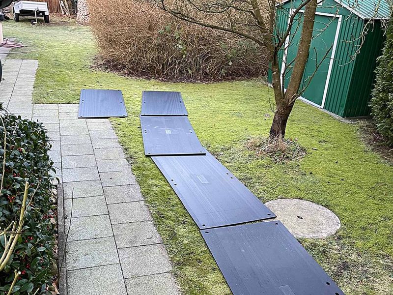 Zum Schutz des Rasens in einem Garten wurden Fahrplatten verlegt. Man sieht einen kahlen Baum und ein Gartenhaus.