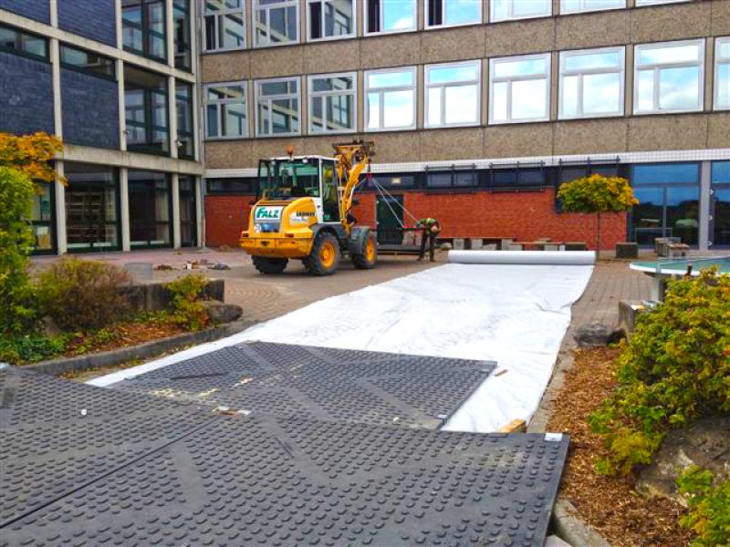 Bagger rollt Geotextil auf Schulgelände aus, um die Arbeitsfläche vorzubereiten, bevor die Kunststoffplatten für den Mobilkran verlegt werden.