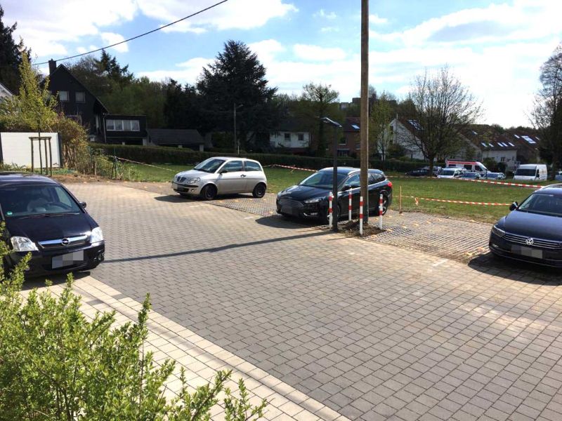 Autos stehen auf einem Parkplatz, der an eine Wiese grenzt. Im Hintergrund sieht man ein Wohngebiet.