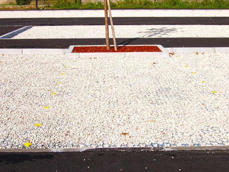 Helle Kiesparkfläche mit Bauminseln und befestigten Stellplätzen für Fahrzeuge.