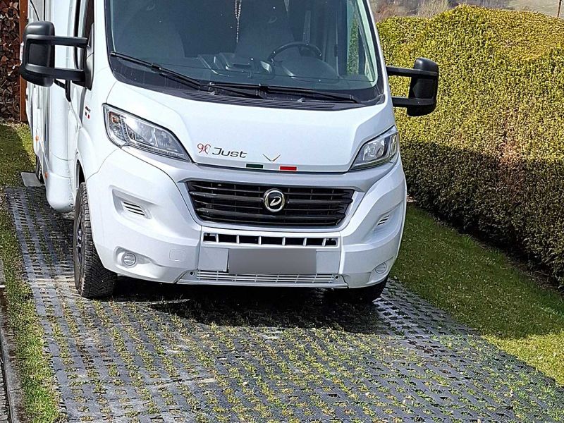 Ein weißes Wohnmobil steht auf einer Fläche aus Kunststoff-Rasengittern. Rechts daneben ist eine dichte Hecke.