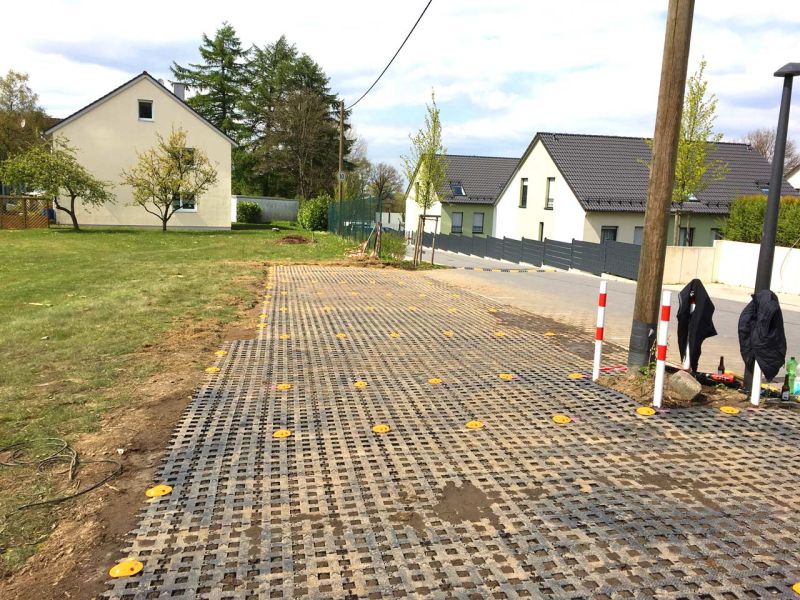 Ein Parkplatz aus Rasengittersteinen neben einer Straße. Auf der anderen Seite ist eine Wiese. Im Hintergrund sieht man Wohnhäuser.