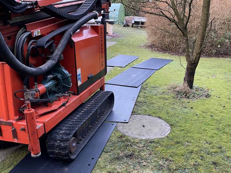 Eine Bohrraupe fährt über Fahrplatten, die auf dem Rasen liegen. Daneben steht ein kahler Baum.