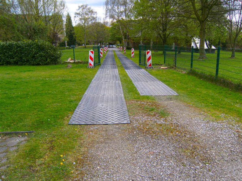 Breiter provisorischer Fahrweg mit Platten über Grünfläche.