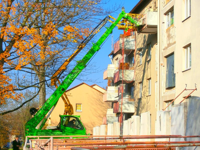 Teleskoplader mit langem Ausleger arbeitet an einem oberen Balkon eines Wohnhauses im begrenzten Platz des Grünbereichs.