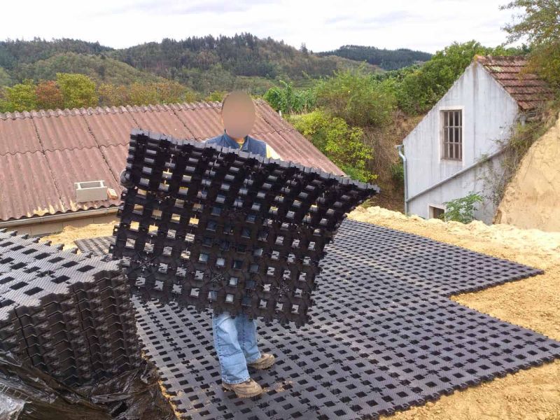 Eine Person verlegt schwarze Kunststoff-Rasengittersteine auf einer vorbereiteten, sandigen Fläche. Sie hält eine flexible Platte hoch, während weitere bereits verlegte Platten den Boden bedecken. Im Hintergrund sind ein Gebäude mit braunem Ziegeldach, ein weißes Haus mit Satteldach sowie eine bewachsene Böschung zu sehen.