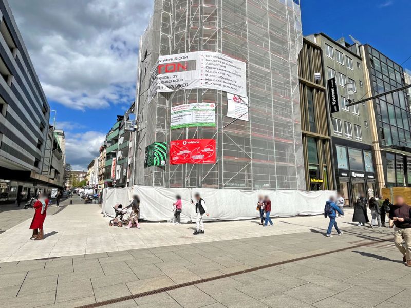 Ein hohes Baugerüst vor einem Gebäude. Passanten laufen davor vorbei.