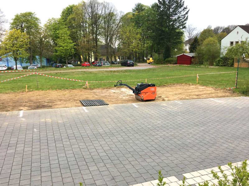 Eine gepflasterte Straße im Vordergrund. Auf einer daran angrenzenden Wiese wurde Rasen entfernt. Ein Rasengitterstein liegt dort.