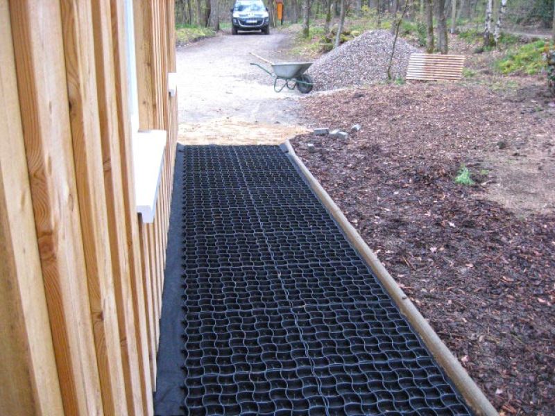 Verlegung eines Kieswegs mit schwarzem Bodengitter entlang eines Holzhauses zur besseren Stabilität und Entwässerung.