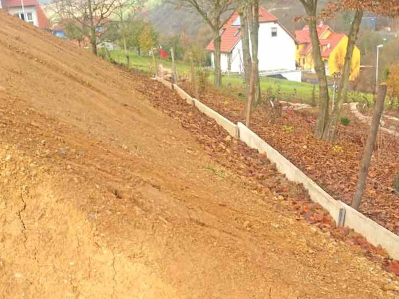 Unbefestigter Hang mit Erdarbeiten und Bretterrand zur späteren Hangstabilisierung. Im Hintergrund sind Bäume und Häuser zu sehen.