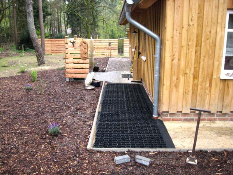 Ein neu angelegter Kiesweg mit einem schwarzen Bodengitter für Stabilität entlang eines Holzhauses.