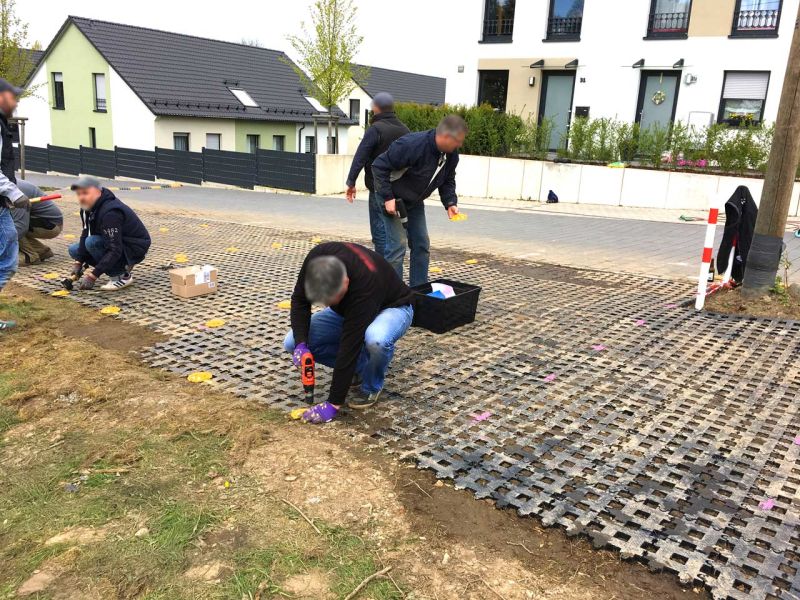 Auf einer Rasenfläche neben einer Straße verlegen mehrere Personen Kunststoff-Rasengittersteine für einen Parkplatz. Im Hintergrund sieht man Wohnhäuser.