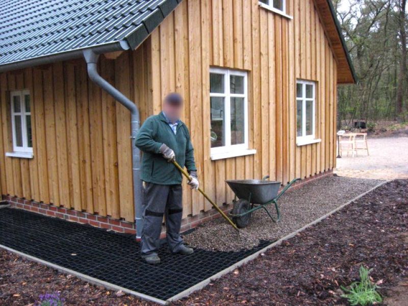 Ein Arbeiter verlegt ein Bodengitter entlang des Hauses, um einen stabilen und wasserfesten Kiesweg anzulegen.