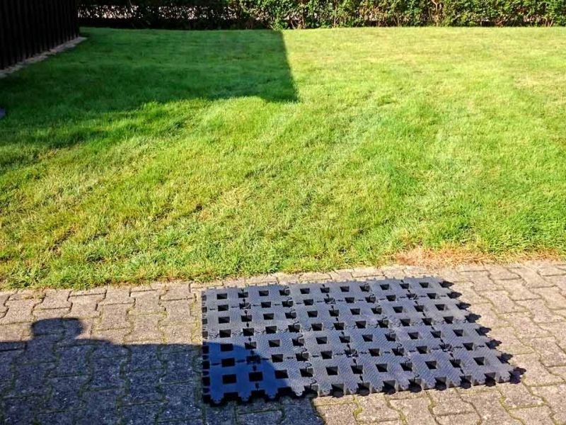 Ein Kunststoff-Rasengitter liegt auf Pflastersteinen neben einer grünen Rasenfläche. Schatten eines Autos sichtbar.