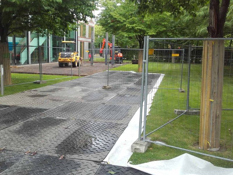 Eine Fläche aus Bodenschutzplatten in einem Park. Bagger stehen darauf. Bauzäune stehen vor den Bäumen.