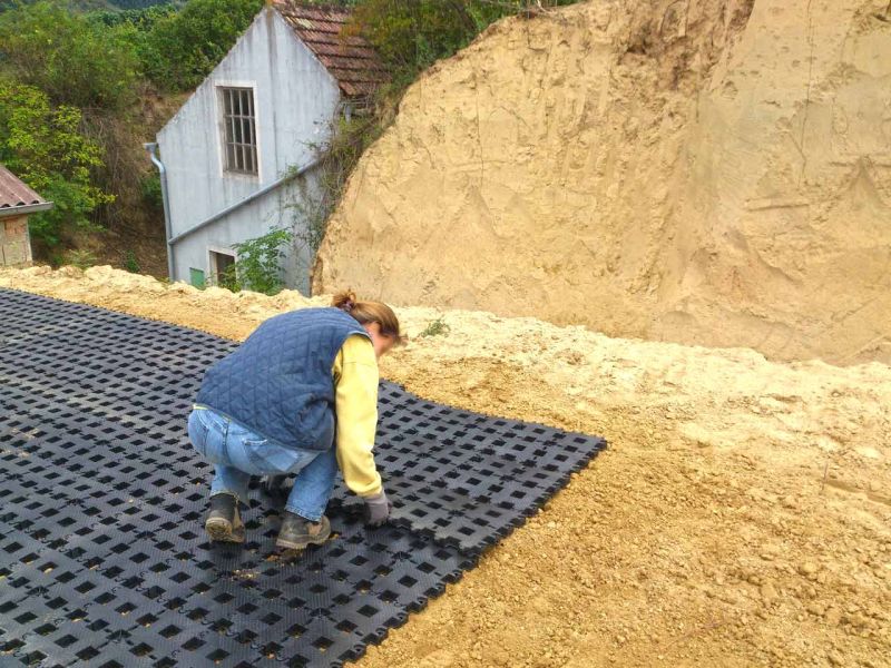 Eine Frau verlegt Rasengittersteine auf einer erdigen Fläche. Im Hintergrund befindet sich ein alt aussehendes Gebäude.