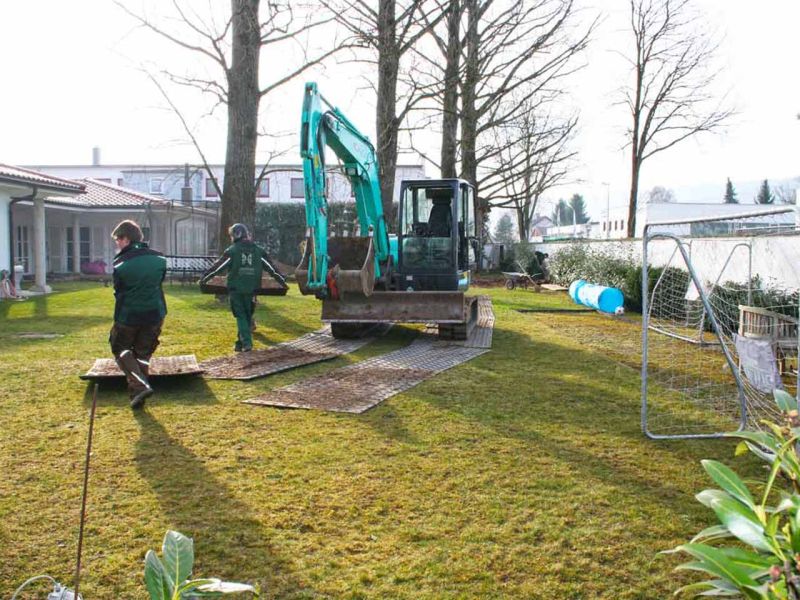 Gartenarbeiter verlegen Fahrplatten, um den Rasen bei Überfahren mit einem Bagger zu schonen.