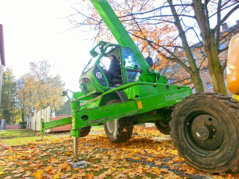 Teleskoplader steht stabil auf ausgefahrenen Stützen im Grünbereich vor Wohnhäusern während der Rückbauarbeiten.