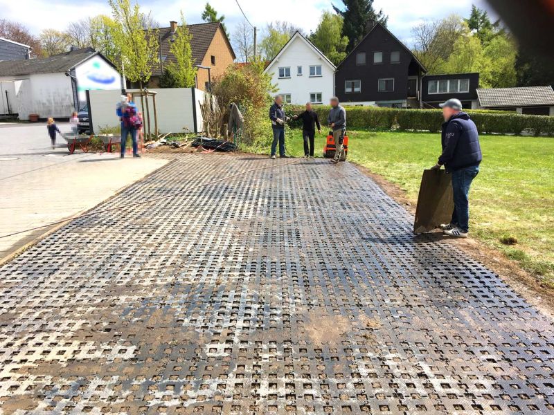 Mehrere Personen verlegen Kunststoff-Rasengittersteine auf einer Rasenfläche neben einer Straße.