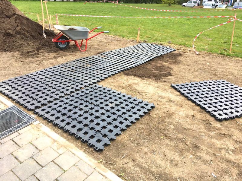 Rasengittersteine aus Kunststoff liegen auf einer Erdfläche. Links daneben sieht man eine Schubkarre, Schippen und einen Erdhaufen.