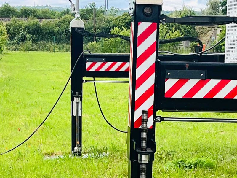 Die Stützen eines mobilen Funkmasts, der auf einer Wiese aufgestellt ist. Im Hintergrund sieht man eine dichte Reihe aus Bäumen. Auf den Stützen sind rot-weiße Markierungen.