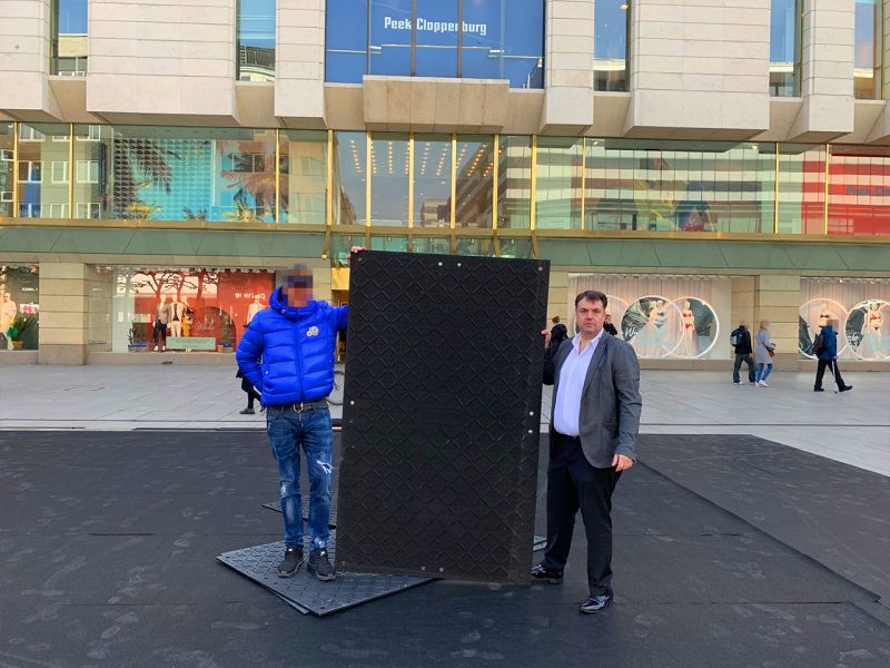 Zwei Personen halten die x:tek46 Bodenschutzplatte aufrecht, um sie für das Foto zu präsentieren. Im Hintergrund ein großes Gebäude mit vielen Schaufenstern.