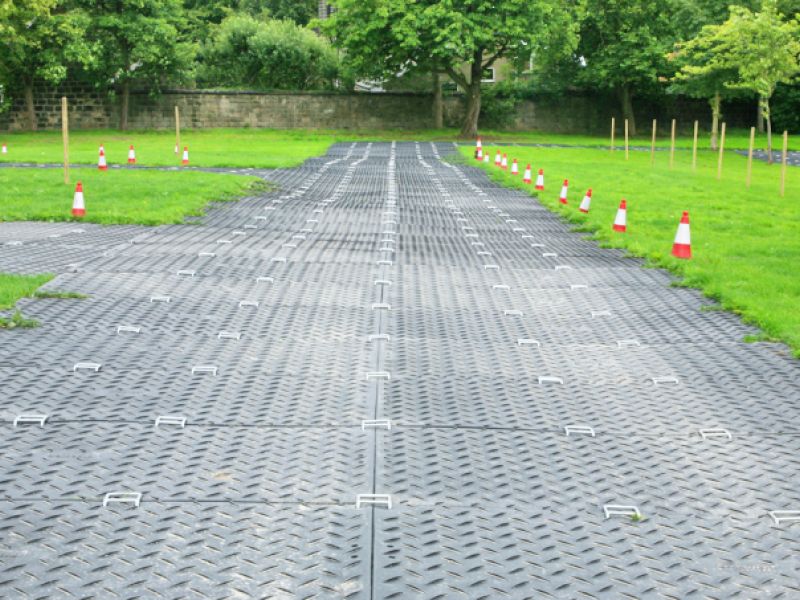 Befestigte Fahrspur aus Platten über eine Wiese mit Absperrkegeln und Bäumen im Hintergrund.