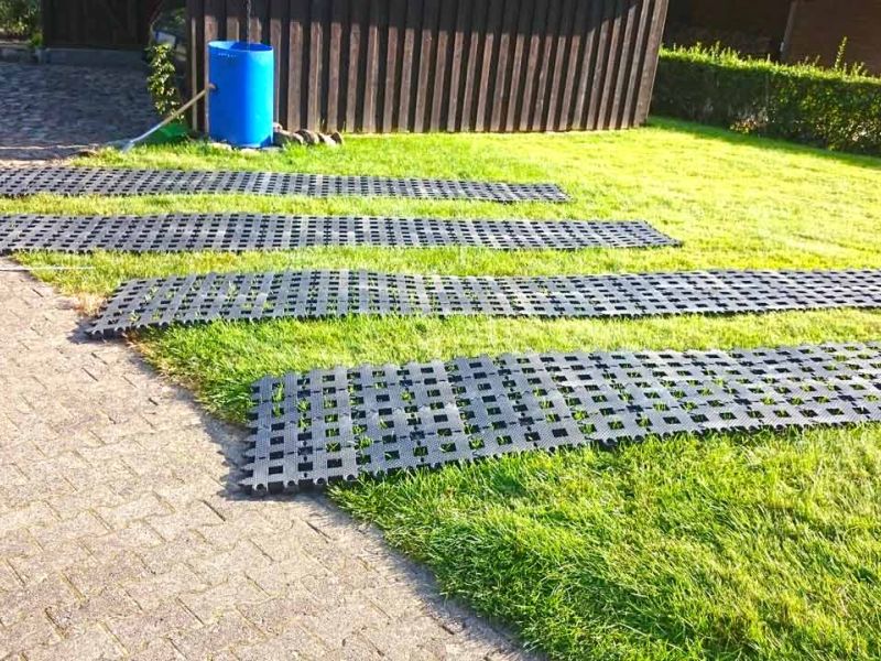 Mehrere Bahnen Rasengittersteine aus Kunststoff liegen auf dem Rasen. Ein blaues Fass steht neben einer Hütte.