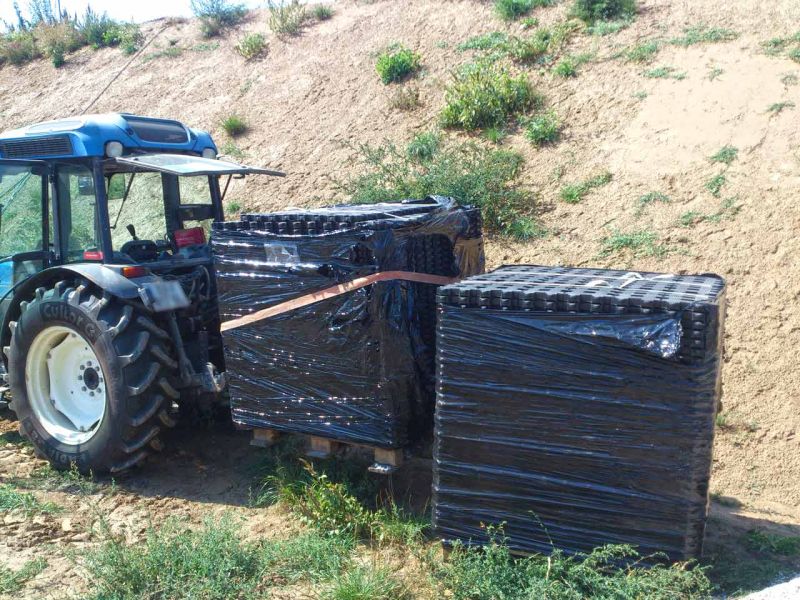 Ein Traktor transportiert Paletten, auf denen Rasengittersteine RGS35 gelagert sind. Im Hintergrund ist ein sandiger Hügel.