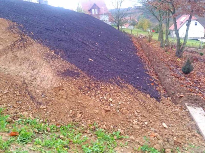 Auf einer erdigen Böschung wurde Mulch verteilt. Im Hintergrund sieht man Wohnhäuser und Bäume.