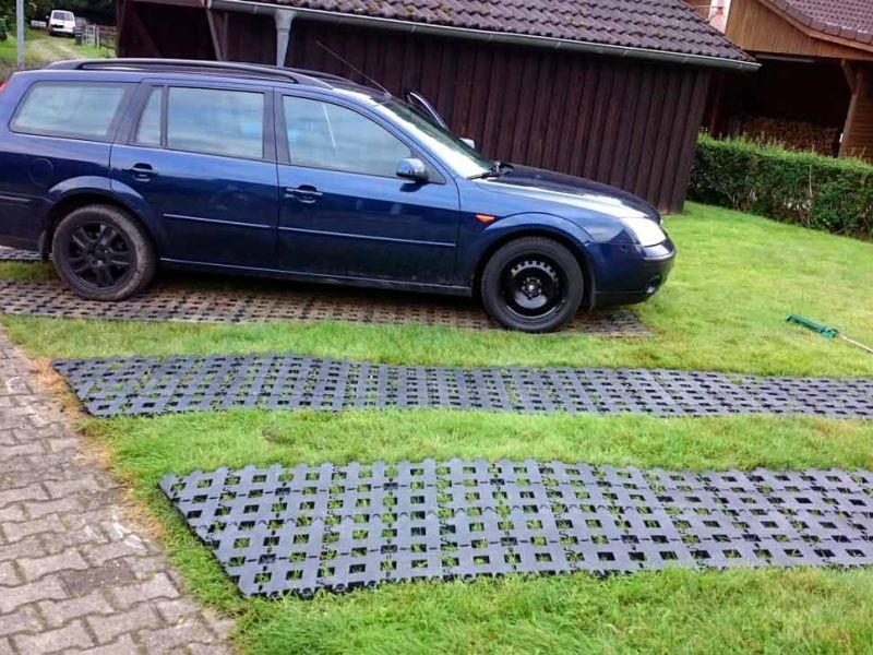 Ein dunkler Kombi parkt auf Kunststoff-Rasengittern. Weitere Platten liegen daneben. Holzhaus im Hintergrund.
