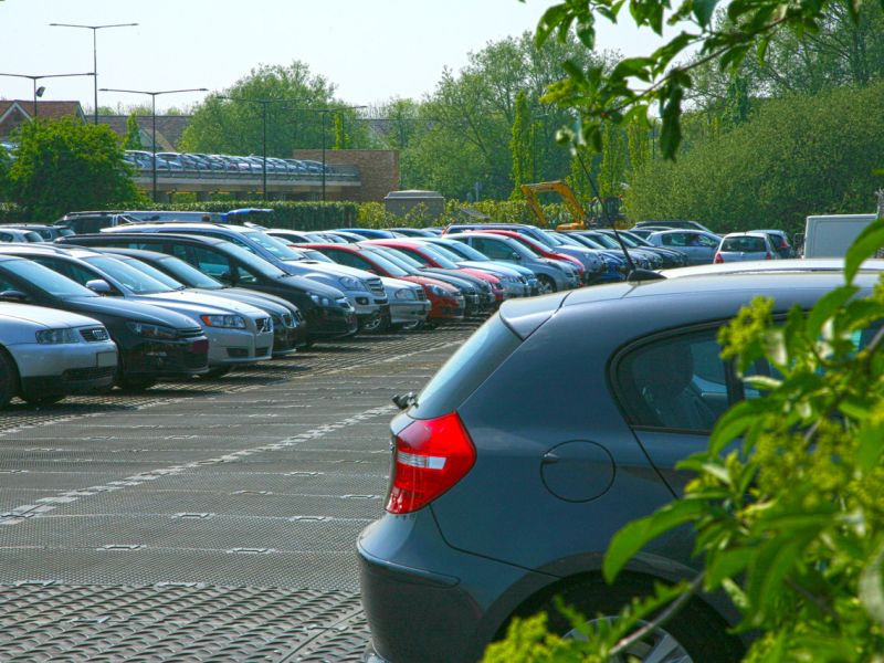 Mehrere geparkte Autos auf einem mit Bodenschutzplatten befestigten Parkplatz, umgeben von Bäumen und Grünflächen an einem sonnigen Tag.