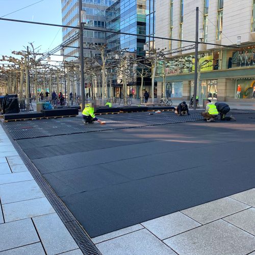 Auf einer Pflasterfläche in einer Fußgängerzone ist eine Gummimatte ausgebreitet. Personen verlegen darauf Bodenschutzplatten.